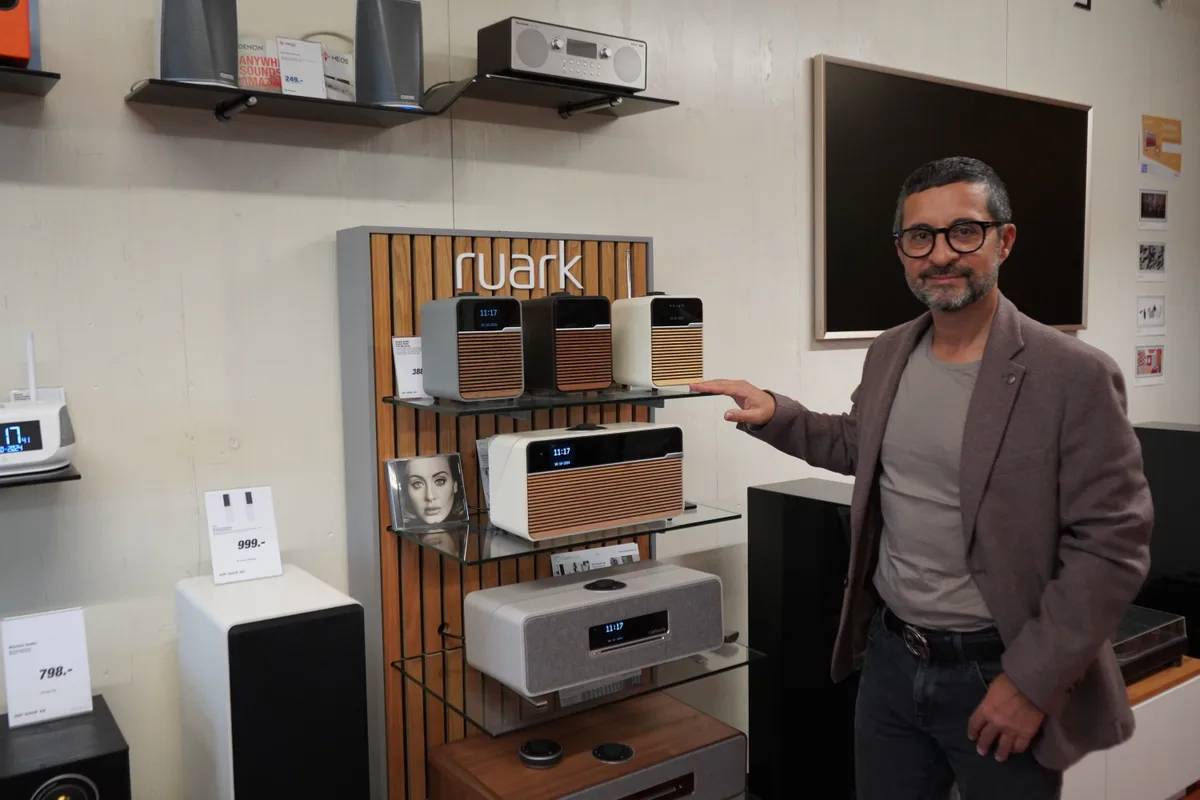 Ein Mann mit Brille in dunklem Sakko steht vor einem Regal mit Radios.