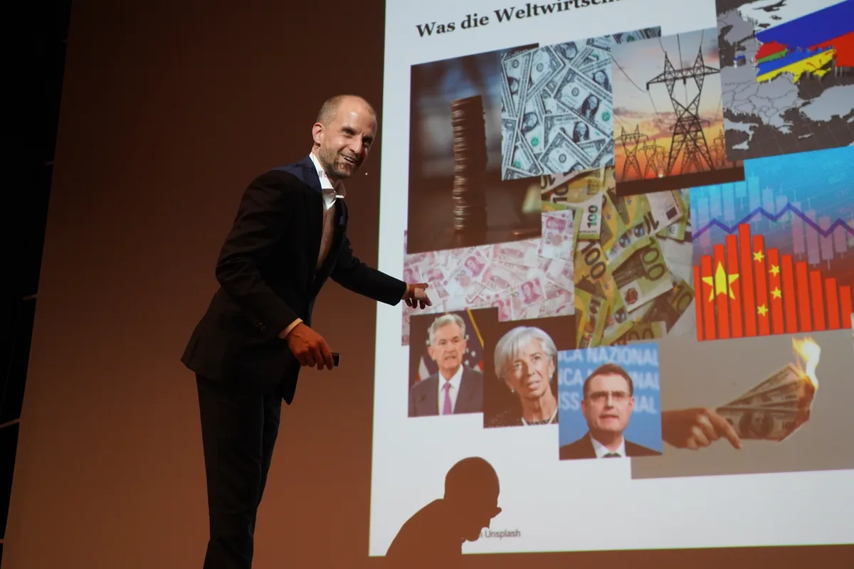 Reto Cueni, Chefökonom der Bank Vontobel, steht auf der Bühne und erklärt einen Slide mit vielen Bildern, der auf die Leinwand projiziert ist.