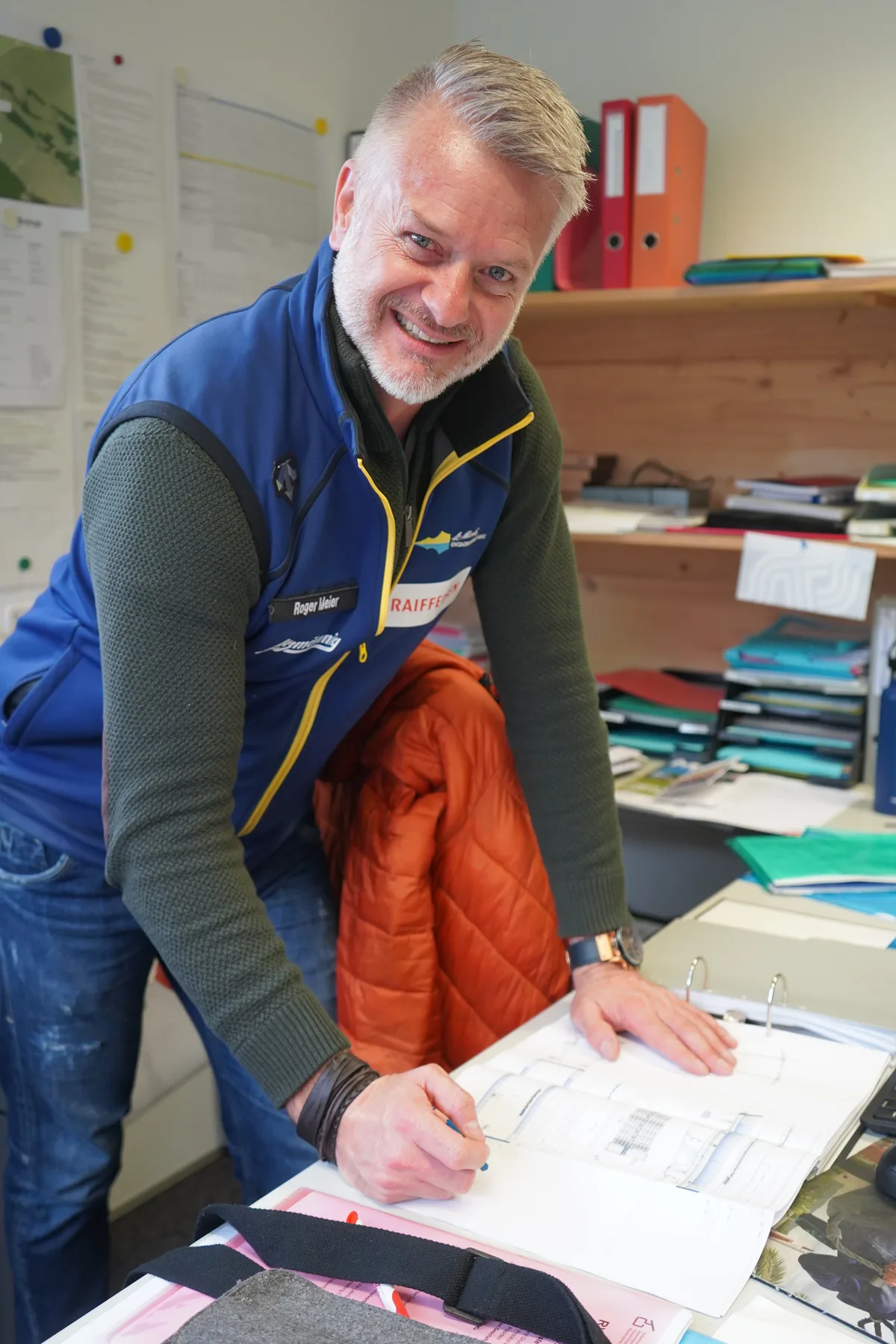 Ein Mann mit Kugelschreiber, über einen Plan gebeugt, lächelt in die Kamera.