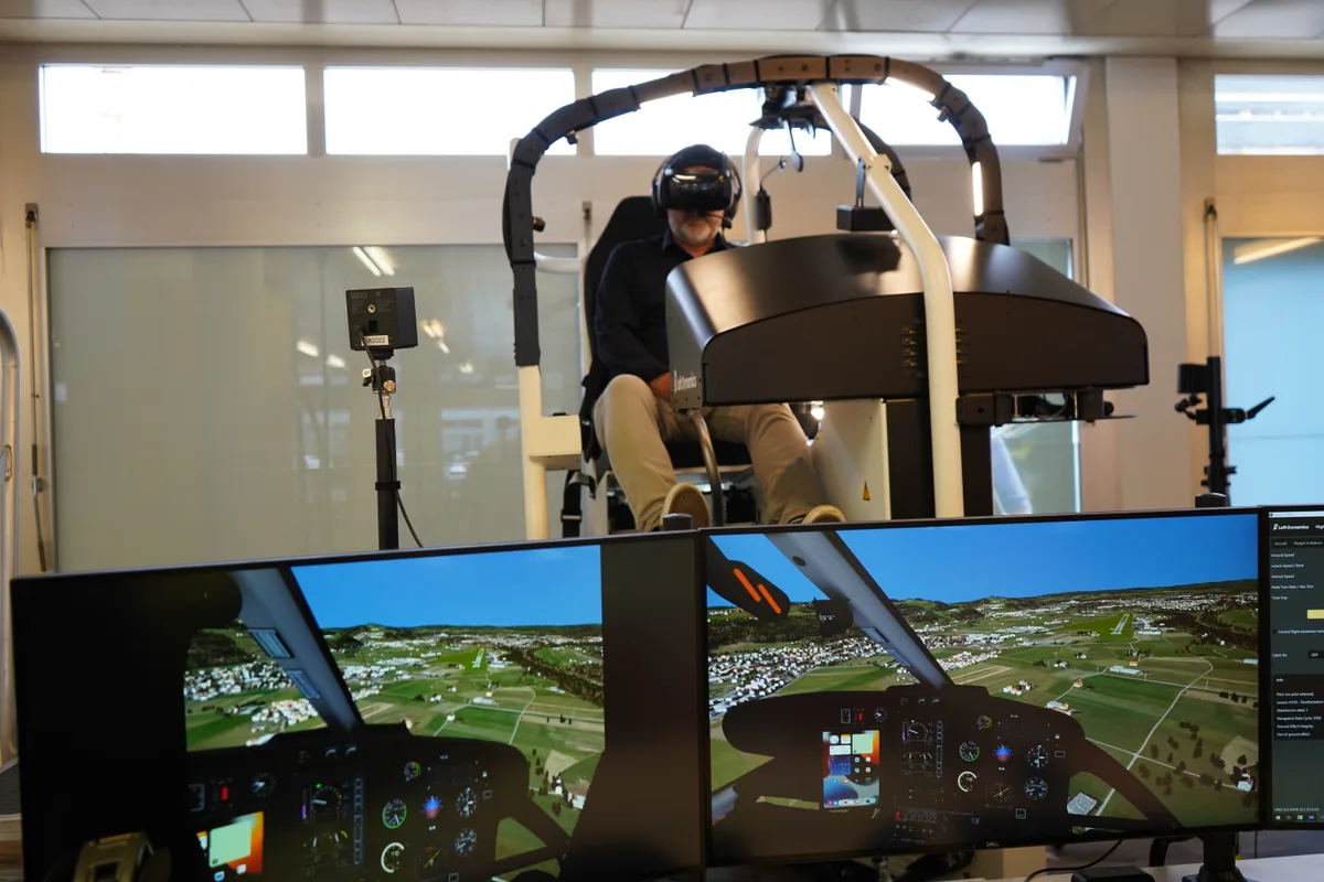 Ein Mann sitzt in einem Flugsimulator. Im Vordergrund zwei Monitore, die zeigen, was der Mann in seiner VR-Brille sieht.