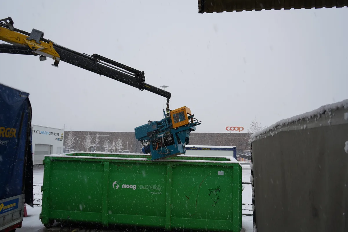 Ein Kran lädt eine Maschine in eine Mulde.