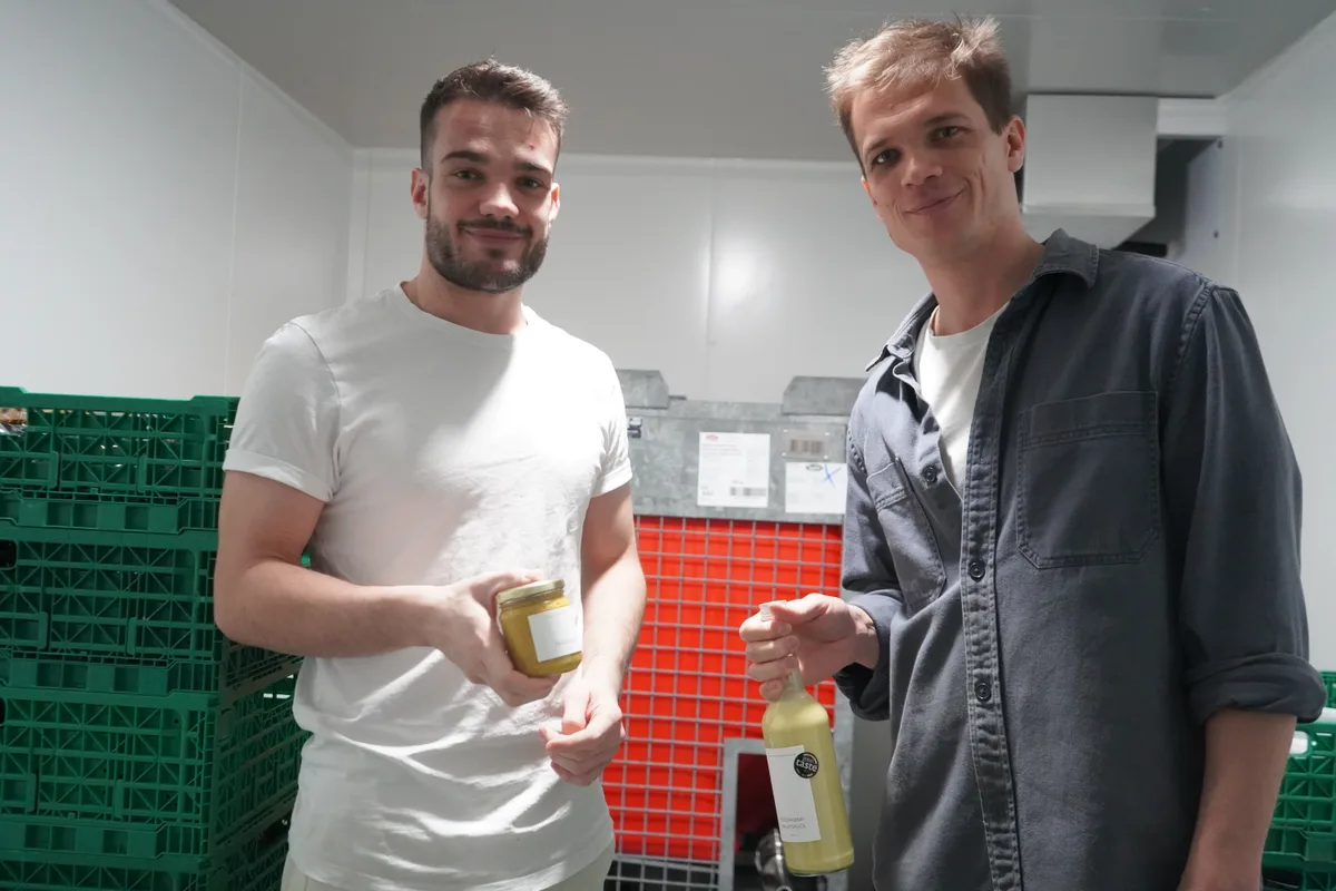Zwei Männer in einem Kühlraum. Einer hält ein Einmachglas, der andere eine Flasche in der Hand.