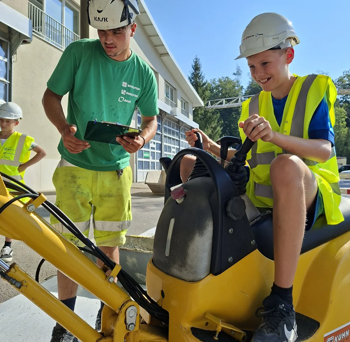 Beim Baggerparcours auf Zeit ging es um Konzentration, Geschicklichkeit und ein Gefühl für Maschinen. Ein Junge mit gelber Warnweste und weissem Helm sitzt auf einem kleinen, gelben Bagger. Er strahlt übers ganze Gesicht.