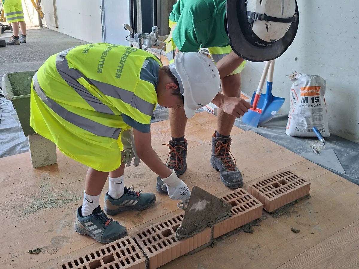 Ganz schön knifflig: Unterstützt von einem Lernenden, darf dieser Junge eine Mauer setzen. Ein Junge mit gelber Warnweste und weissen Helm streicht mit einer Maurerkelle Mörtel auf einen Backstein. Daneben steht ein Erwachsener und leitet ihn an.