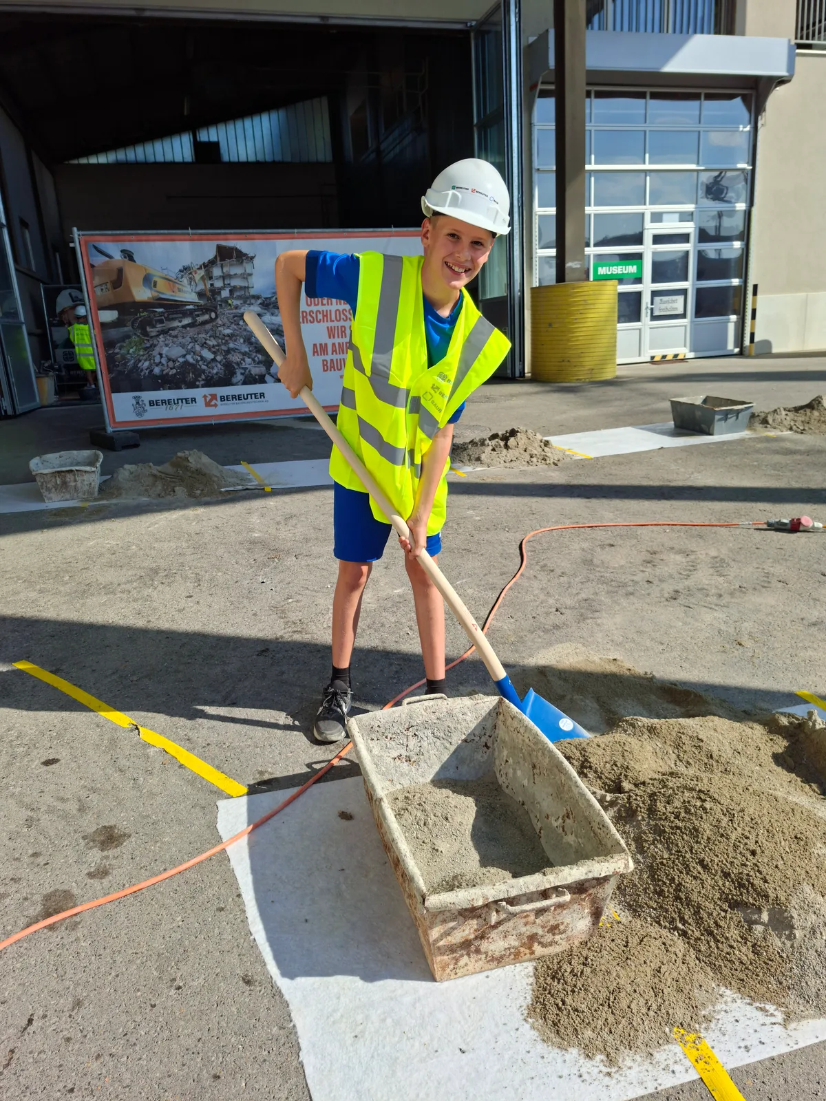 Im Zentrum steht der Spass. Ein Junge schaufelt Sand aus einem Behälter. Er trägt eine gelbe Warnweste und einen weissen Helm.