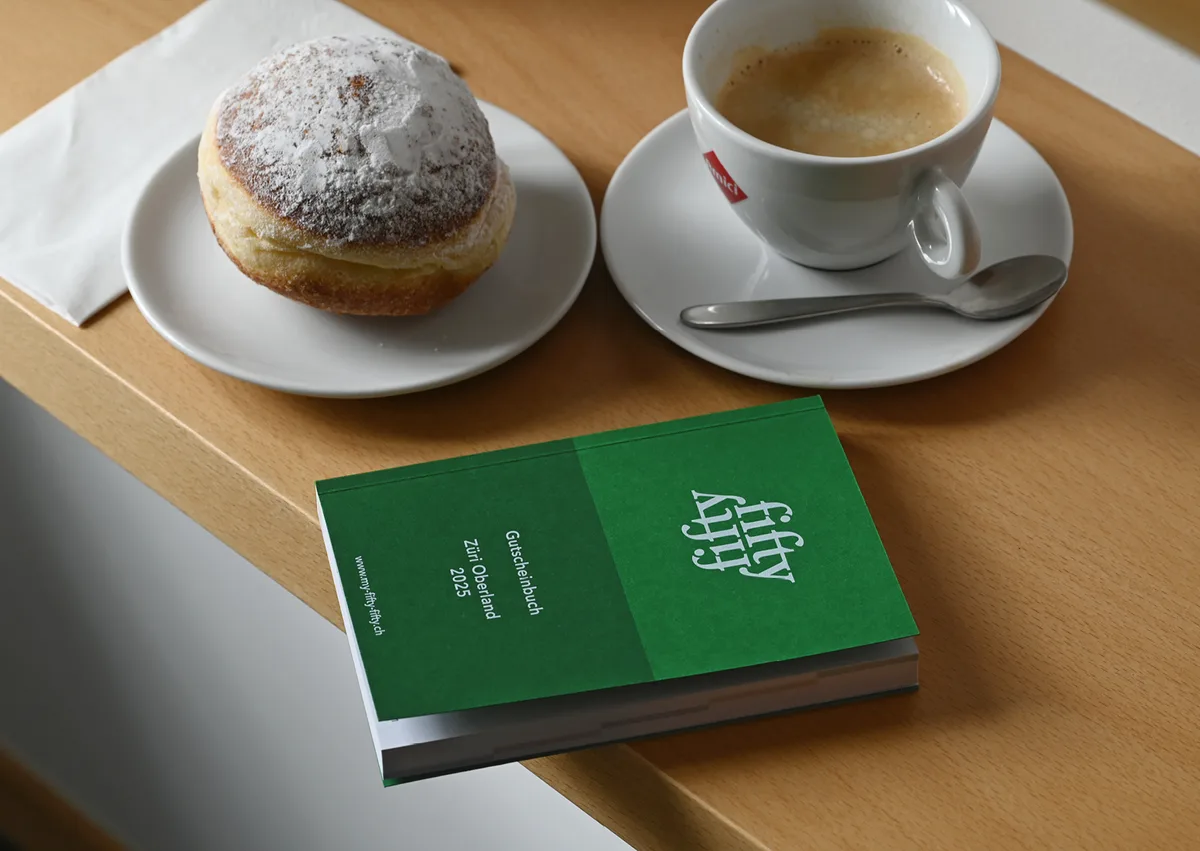 Ein grünes Gutscheinbuch mit dem Titel «fifty-fifty» liegt auf einem Tisch. Daneben ein Kaffee und ein Berliner.