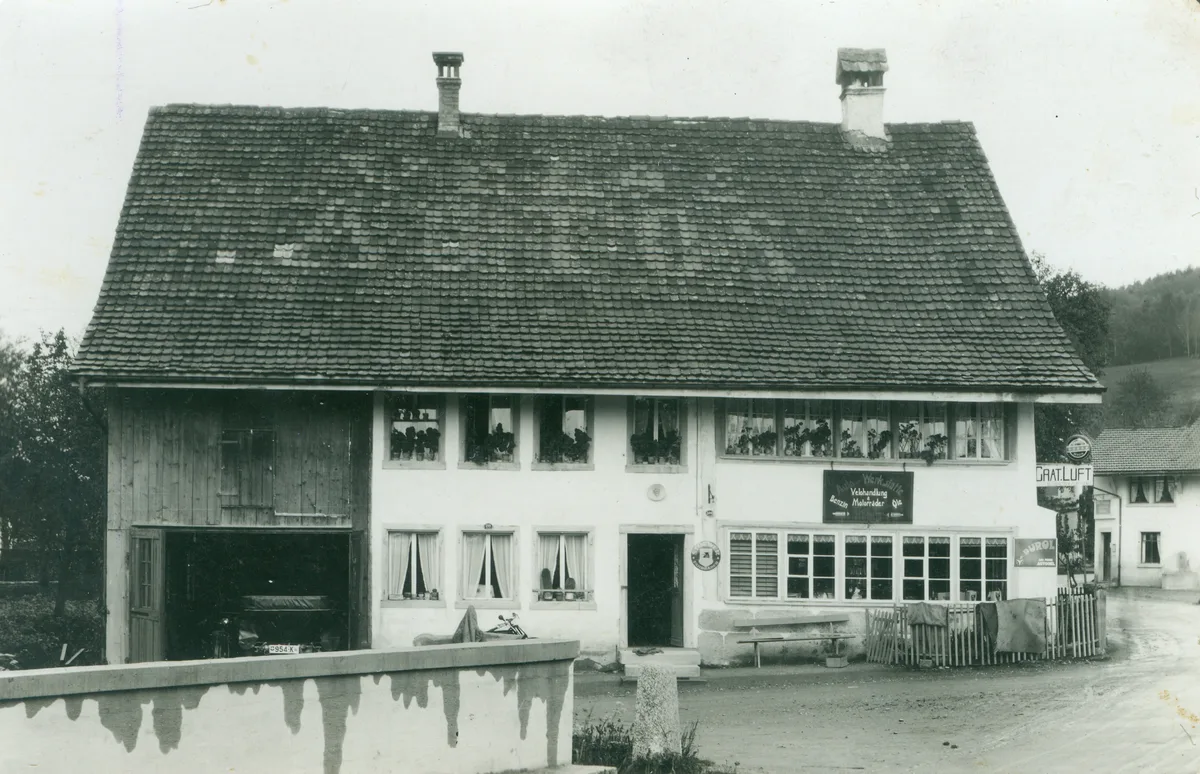 Ein altes Bauernhaus, das als Autowerkstatt dient. Schwarz-Weiss-Aufnahme.