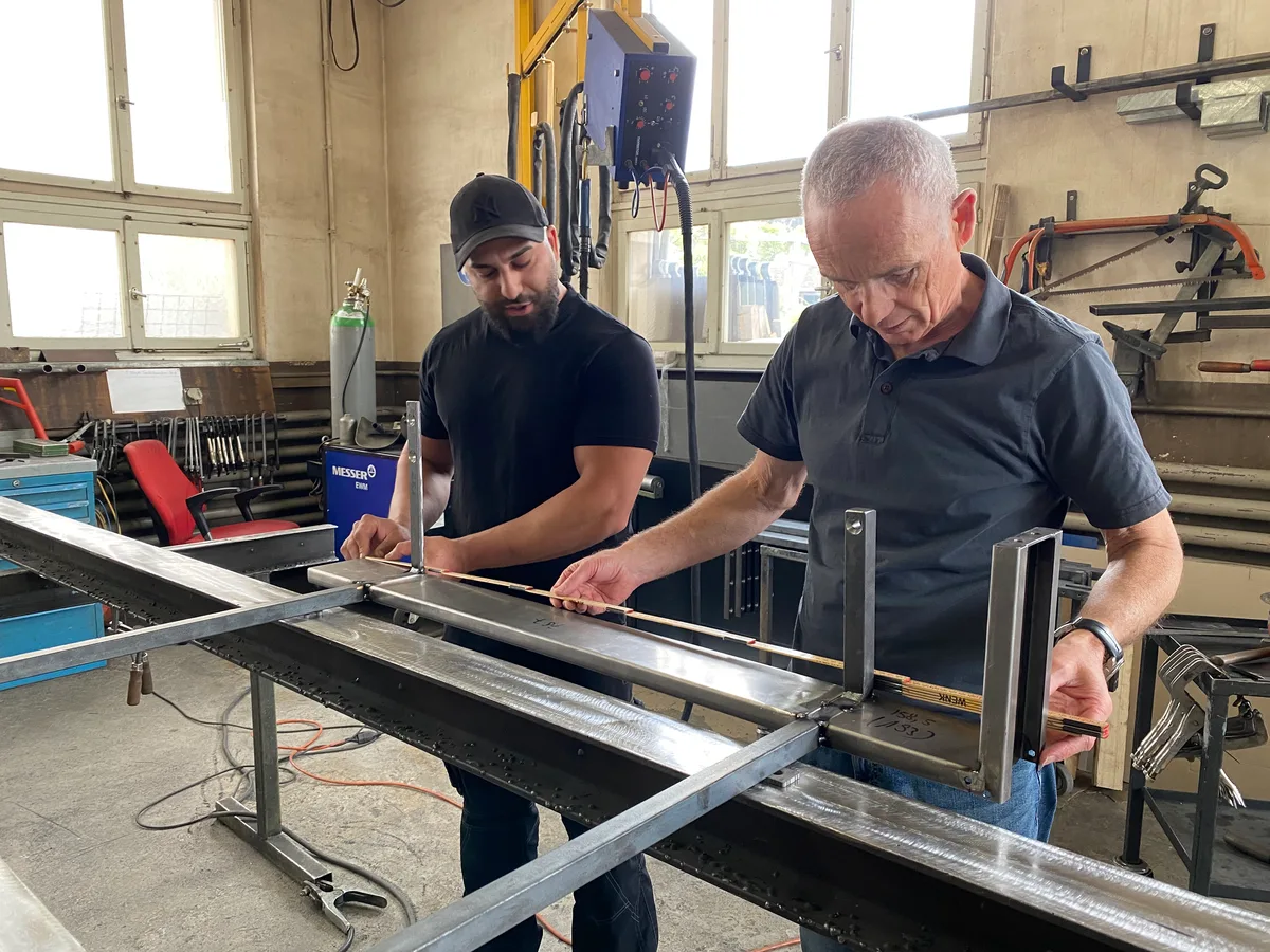 Zwei Männer stehen in einer Schlosserwerkstatt und messen eine Metallkonstruktion aus.
