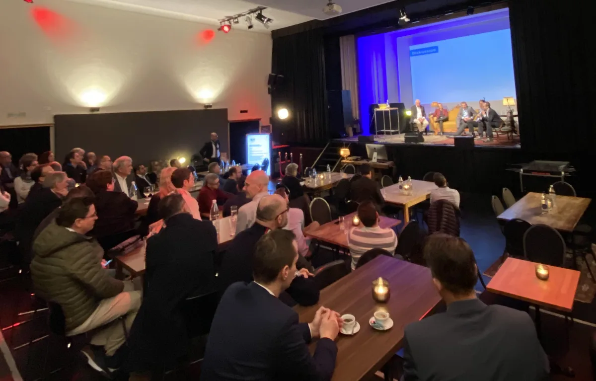 Leute im Saal folgen einer Podiumsdiskussion. Auf der Bühne sitzen vier Männer und eine Frau.