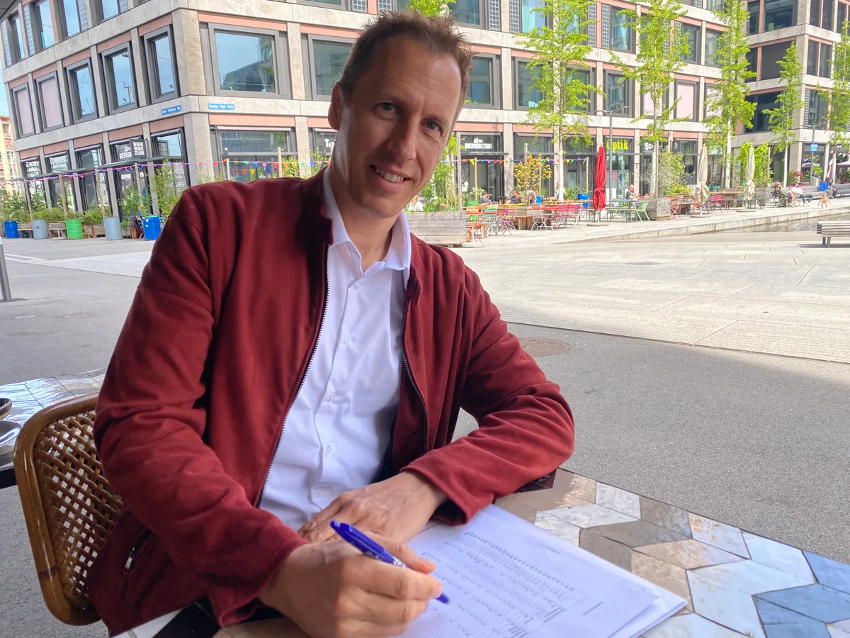 Ein Mann in roter Wildlederjacke sitzt an einem Tisch. Vor sich hat er Dokumente, in der linken Hand hält er einen Kugelschreiber.