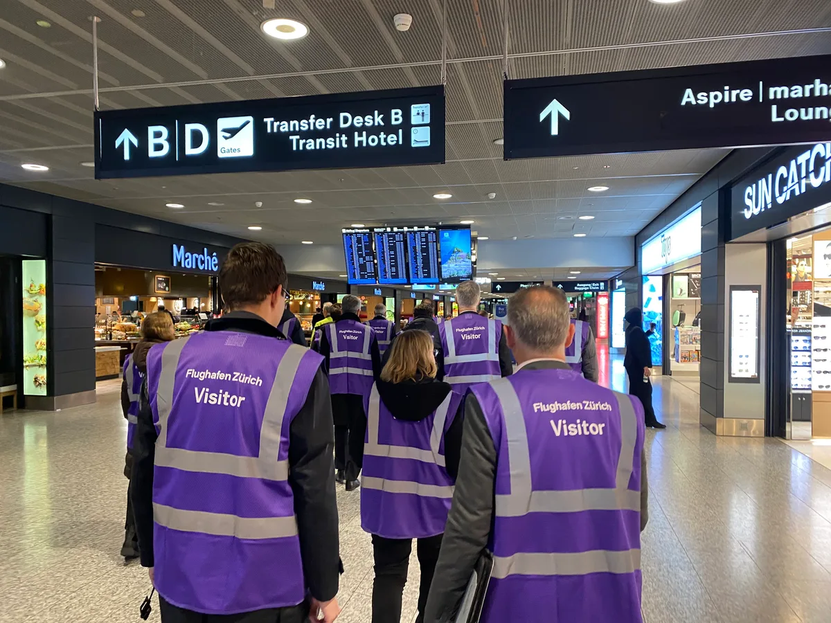 Eine Gruppe von Menschen am Flughafen Zürich. Sie tragen violette Westen, auf denen Visitor steht.