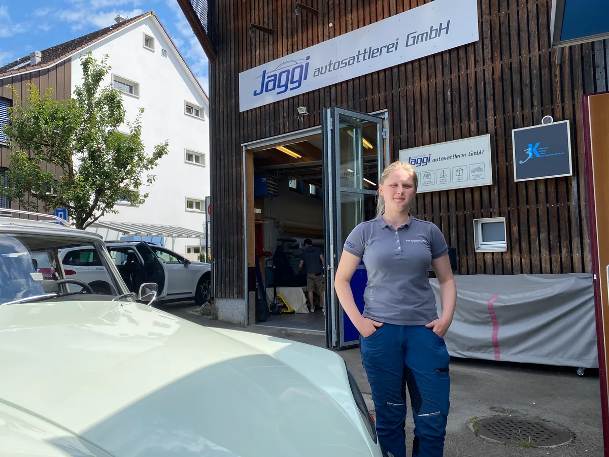 Eine junge Frau steht vor einer Werkstatt. Das Haus ist mit Jaggi Autosattlerei GmbH angeschrieben.