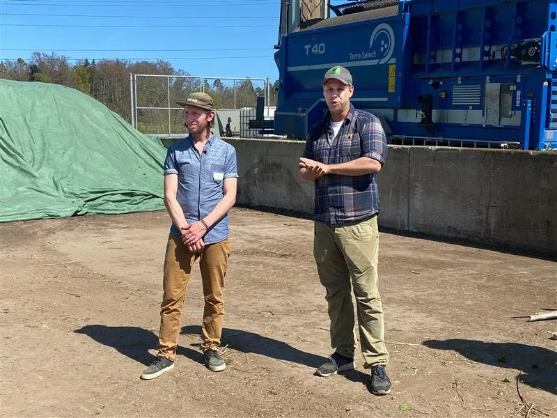 Jojo Casanova von Kompotoi und Michael Kunz, Inhaber der Kunz Baumschulen in Uster. Zwei Männer stehen auf einem Betonplatz und erklären etwas. Beide tragen Baseballmützen und legere Kleidung. In Hintergrund sind eine blaue Maschine und ein mit grüner Folie abgedeckter Haufen zu sehen.