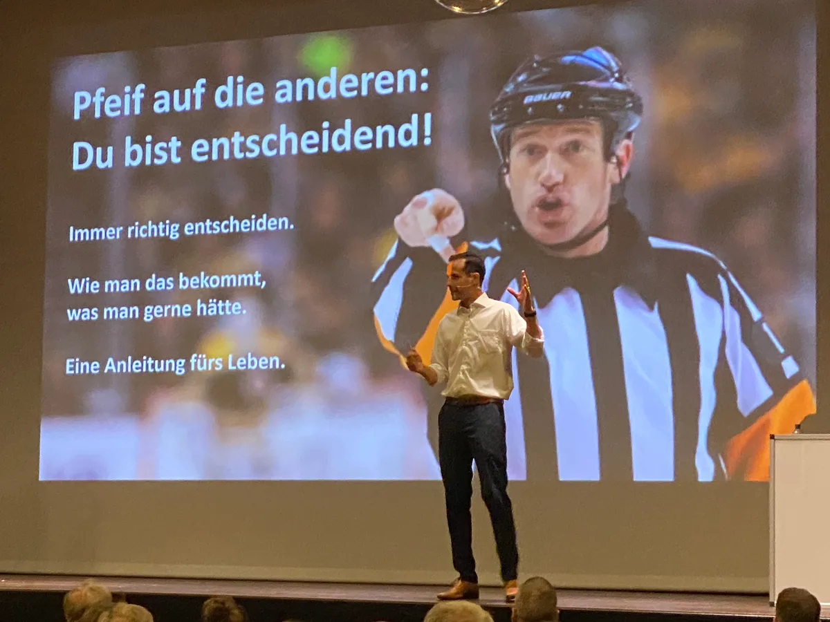 Ein Mann steht auf der Bühne des Ustermer Stadthofsaals und referiert. Im Hintergrund sieht man ein grosses Bild dieses Mannes als Eishockeyschiedsrichter.