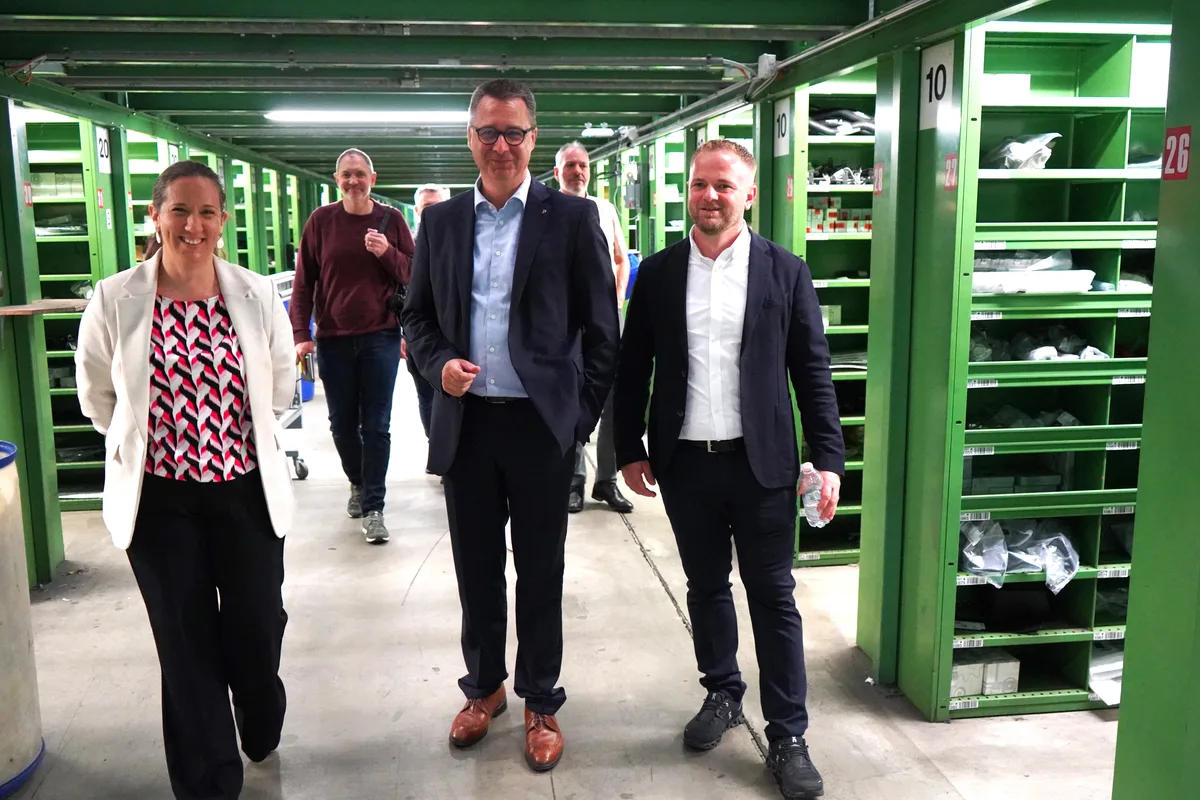 Eine Frau und zwei Männer im Anzug schreiten durch ein Warenlager.