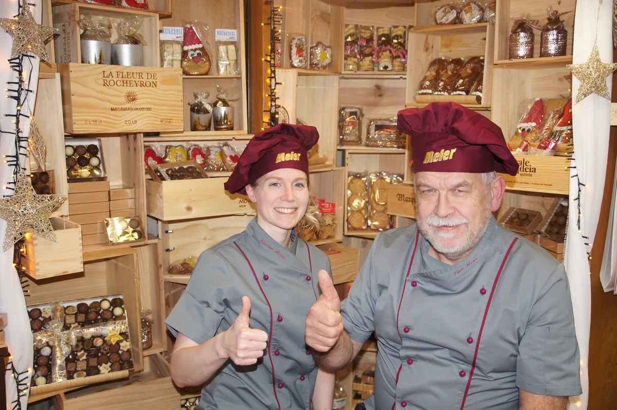 Ein Mann und eine junge Frau in grauen Hemden und bordeauxroten Mützen. Beide halten einen Daumen in die Luft.