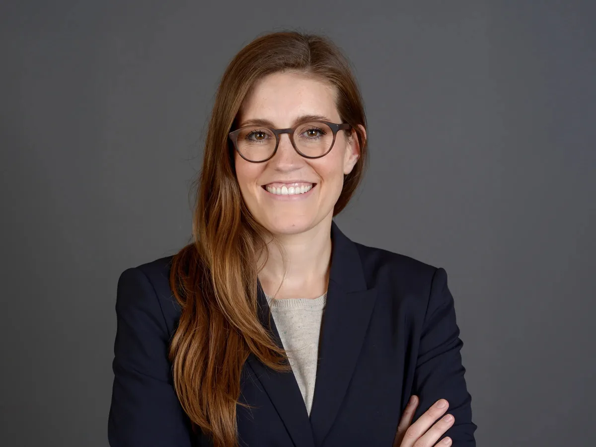 Porträtbild einer Frau mit Brille und langen, braunen Haaren. Sie trägt einen dunklen Blazer.