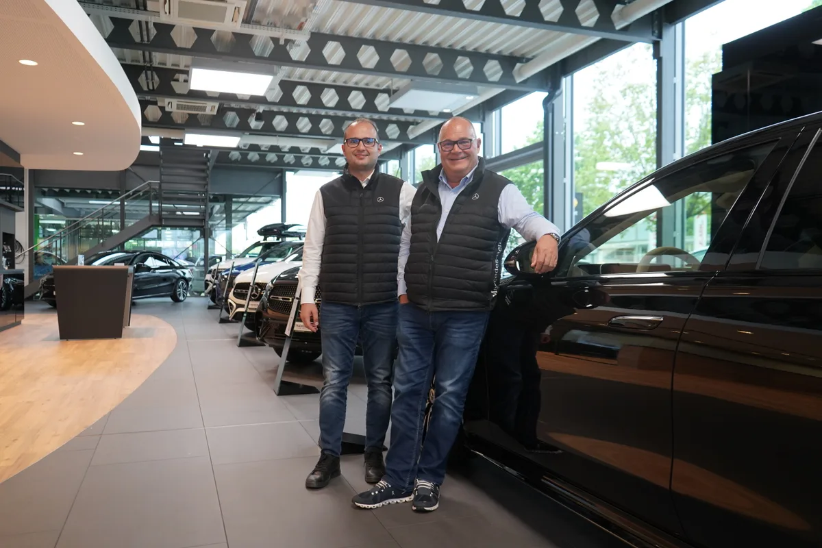 Zwei Männer stehen in einem Showroom für Neuwagen an einen Mercedes gelehnt.