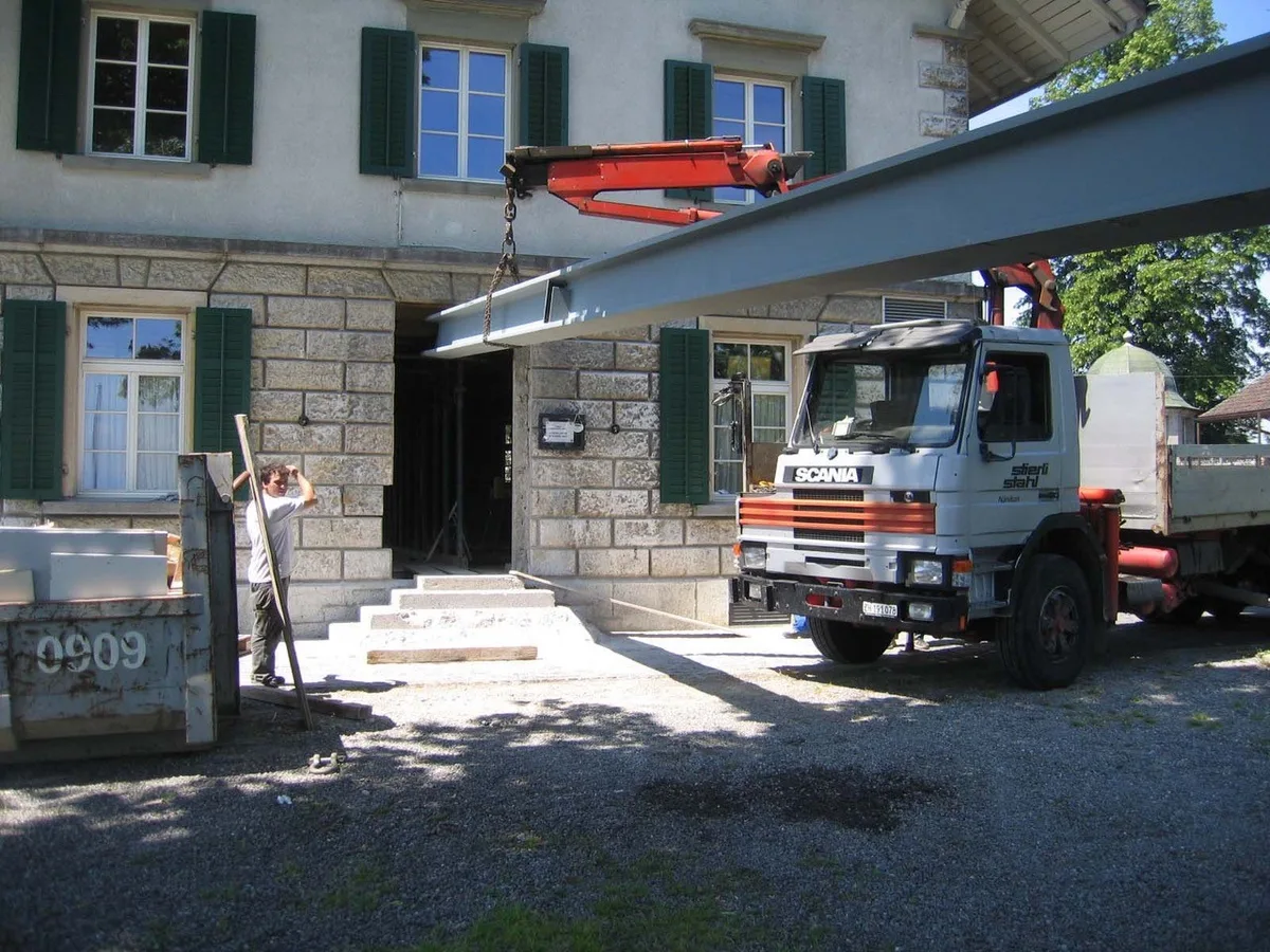 Ein Lastwagen mit Kran hält einen grossen, grauen Eisenträger vor einem historischen Gebäude.
