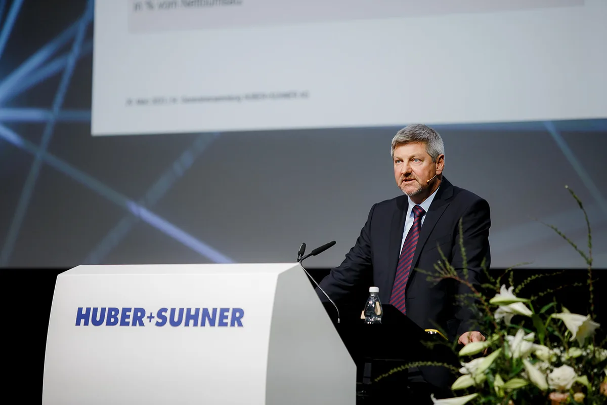 Ein Mann im Anzug steht hinter einem Rednerpult. Dieses ist mit «Huber+Suhner» beschriftet.