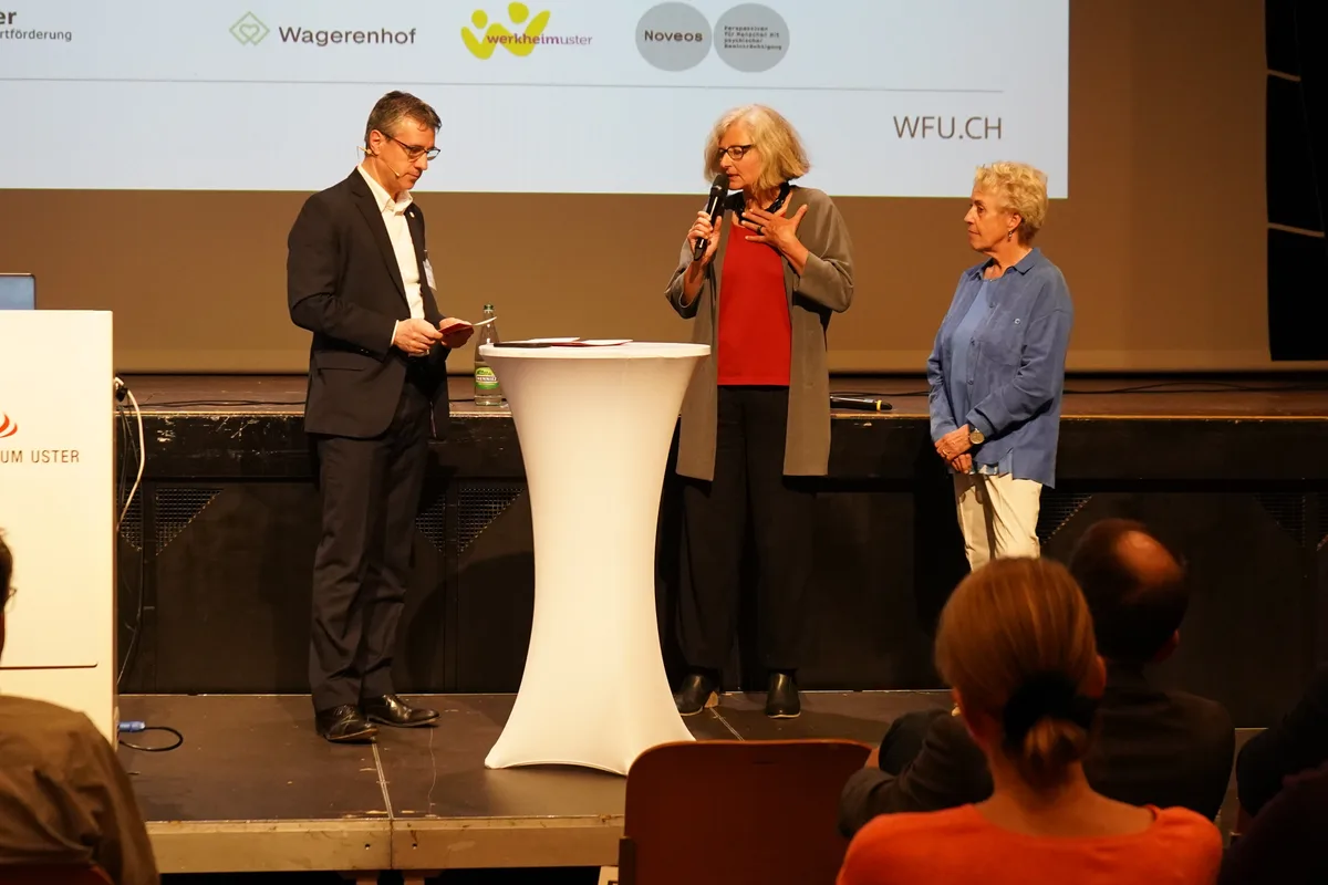 Ein Mann im Anzug steht mit zwei Frauen auf einer Bühne.