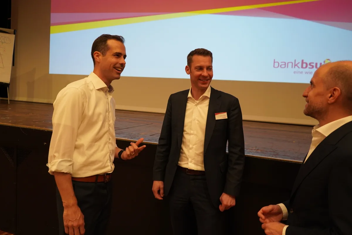 Drei Männer unterhalten sich vor einer Bühne. Im Hintergrund das Logo der Bank BSU.