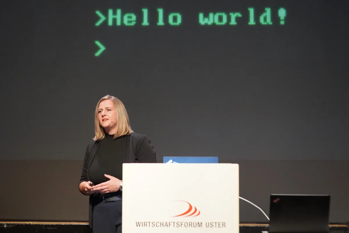 Eine Frau referiert auf einer Bühne. Dahinter sieht man einen Schriftzug «Hello world!».