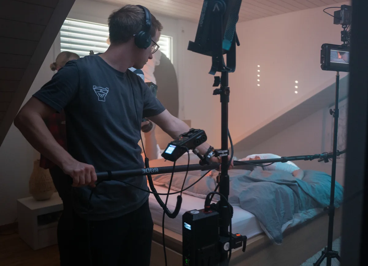 Szene am Filmset, im Vordergrund hält ein Mann mit Kopfhörern ein Mikrofon.