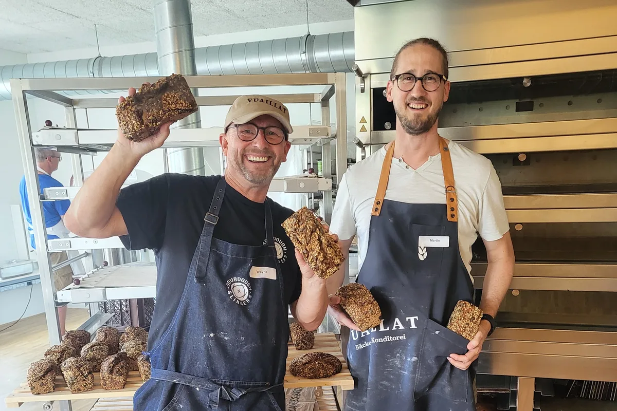 Zwei Männer halten in einer Backstube Brote in ihren Händen und lächeln in die Kamera.