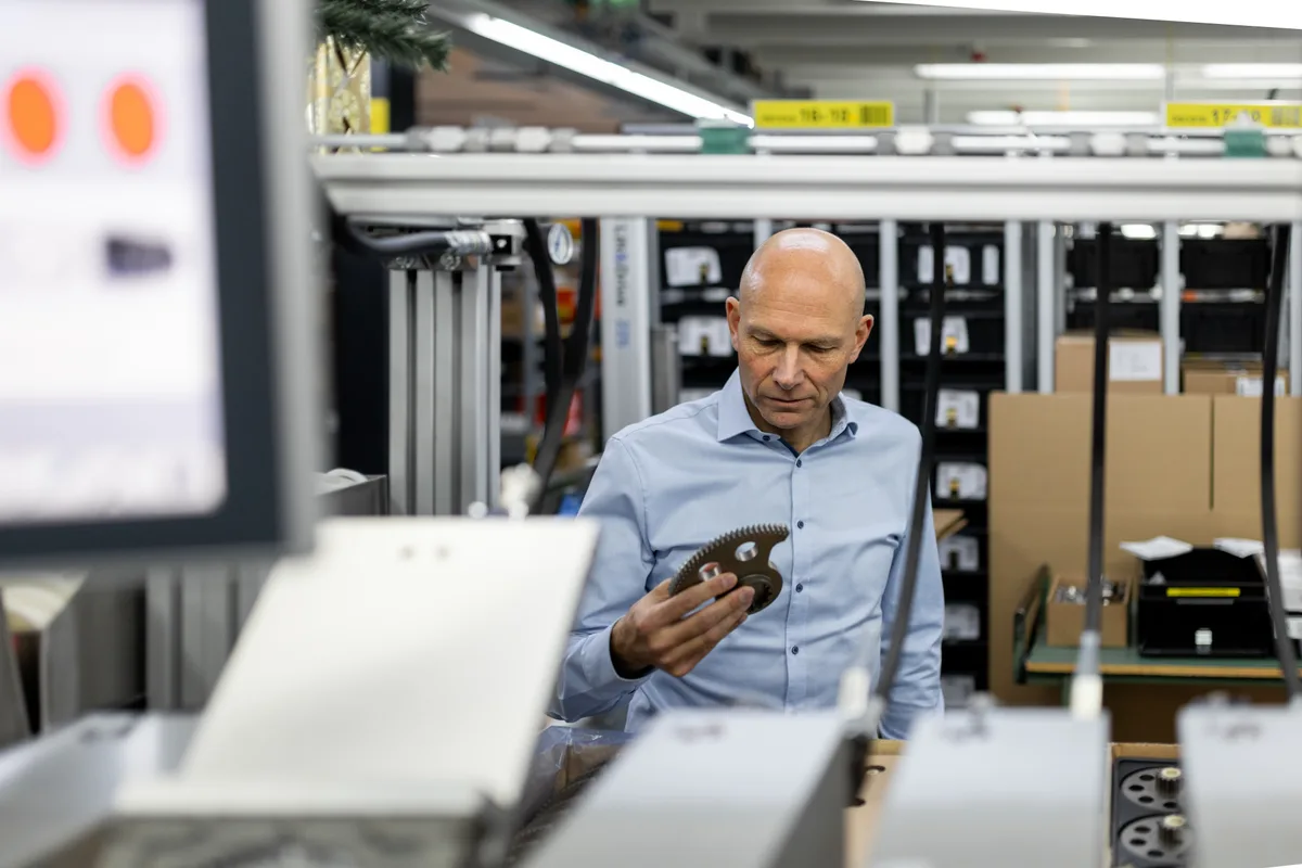 Ein Mann im offenen blauen Hemd hält ein Bauteil in der Hand.