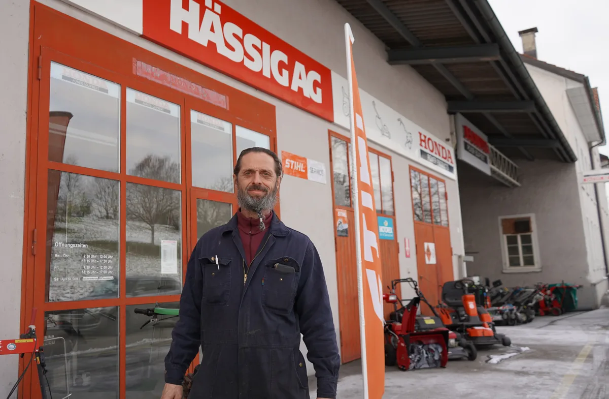 Ein Mann im blauen Arbeitsoverall steht vor einer Geschäftsliegenschaft. Man sieht den Firmennamen Hässig AG und Geräte wie Schneefräsen oder Rasenmäher.