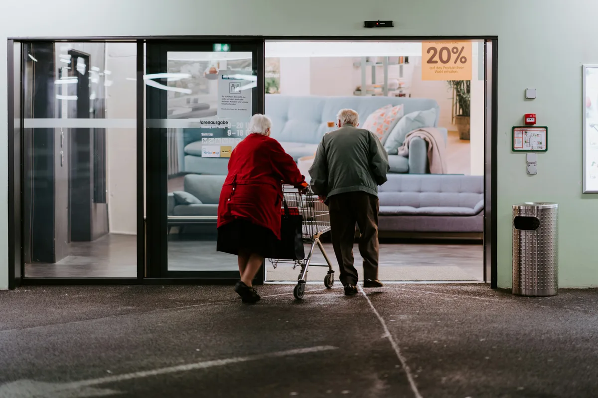 Ein altes Paar – sie mit einem Rollator – verlässt ein Gebäude.