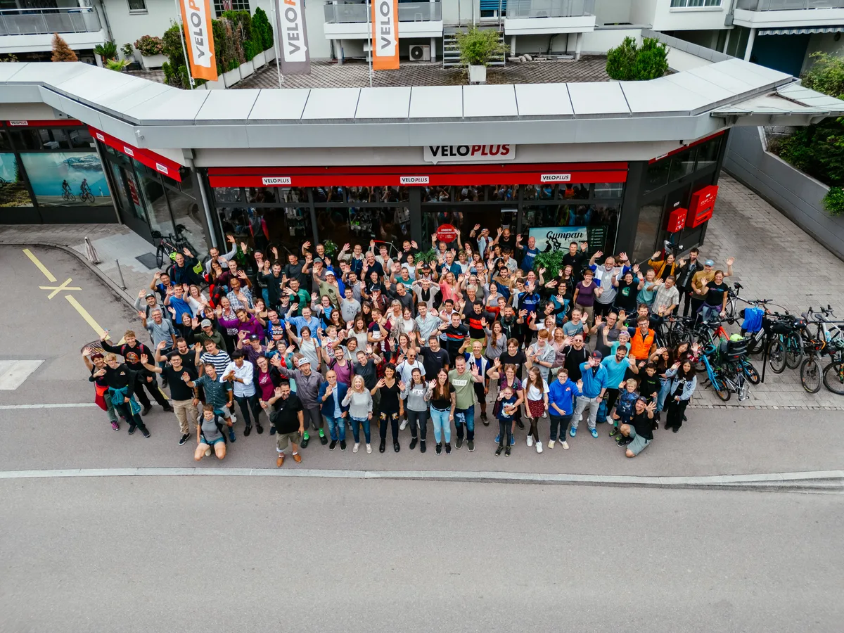 Luftaufnahme: Viele Menschen stehen vor einem Veloplus-Laden und winken in die Kamera.