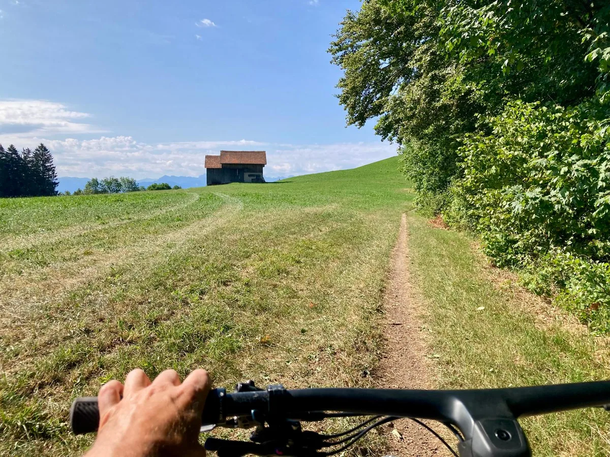 Ein Wanderweg mit einem Stall links. Vorne ist der Lenker eines Mountainbikes.