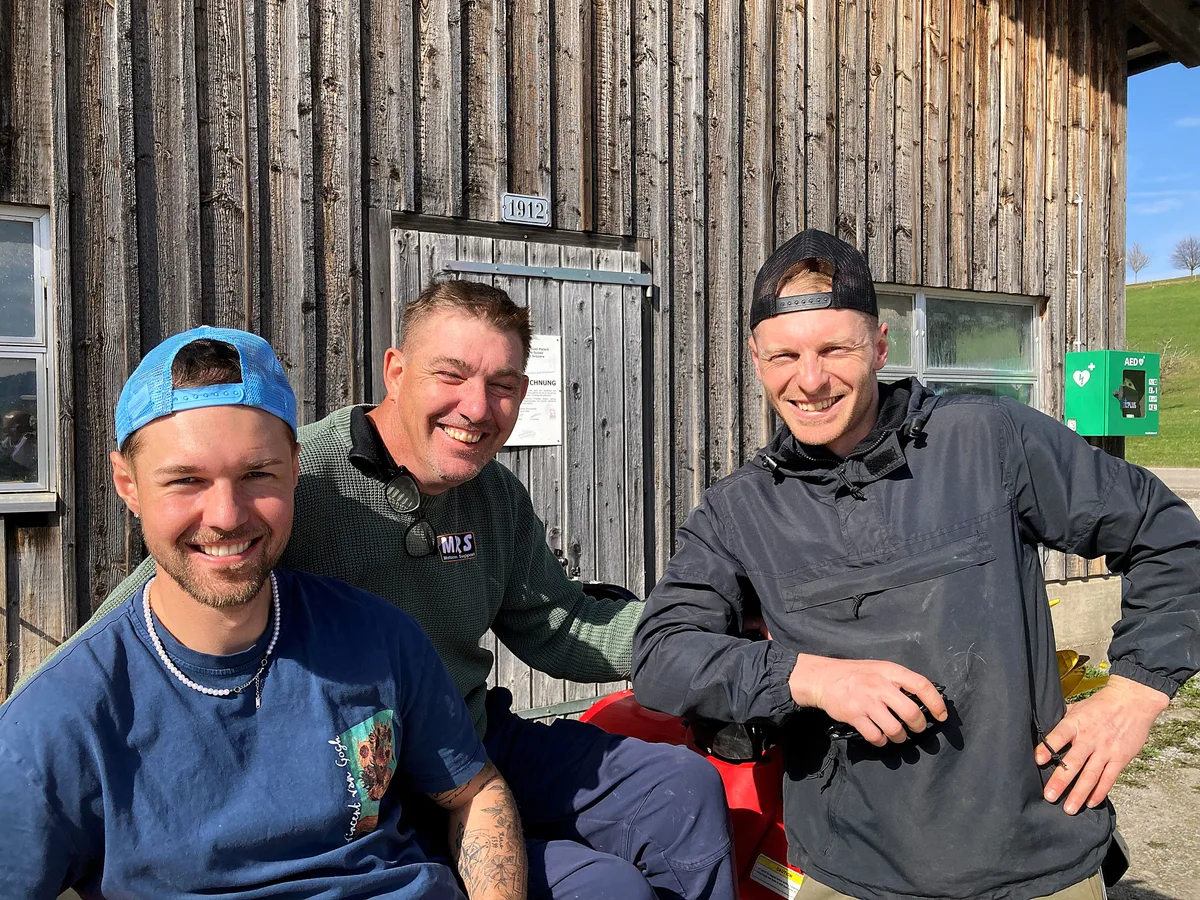 Drei Männer vor einem Stall, zwei davon tragen eine Dächlikappe verkehrt herum.