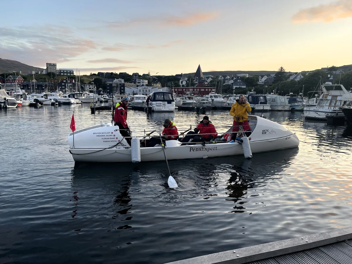 Der Blick aufs Boot lässt gut erahnen, wie wenig Platz die vier Ruderer zur Verfügung hatten. Vier Personen auf einem Ruderboot in einer Bucht.