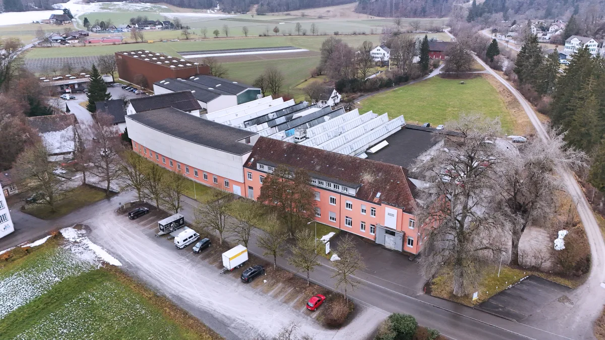 Der Hauptsitz von Kuhn Rikon. Am 16. März 1926 erwarb Heinrich Kuhn das Unternehmen, das heute in vierter Generation von Dorothee Auwärter geführt wird. Luftaufnahme von Kuhn Rikon in Rikon. Man sieht das rosafarbene Verwaltungsgebäude und die Fabrikationshalle.
