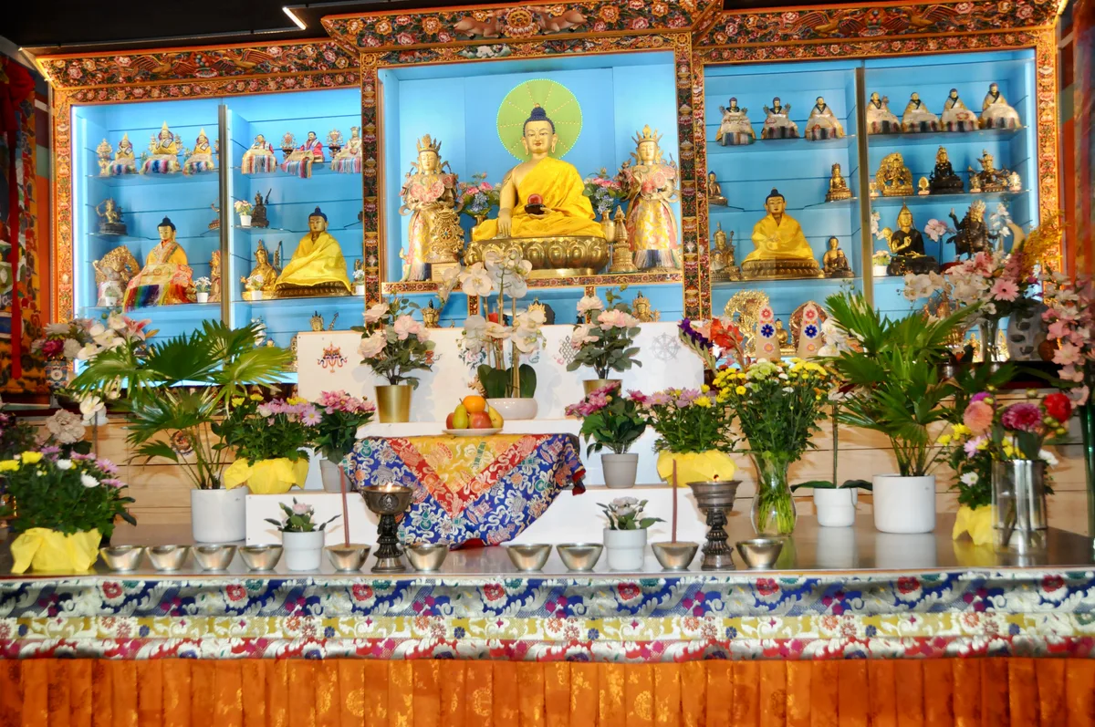 Hinter dem «Blumenmeer» sind die Buddha-Statuen aus den verschiedenen Kulturen zu sehen. Man sieht einen farbenprächtigen Altar.