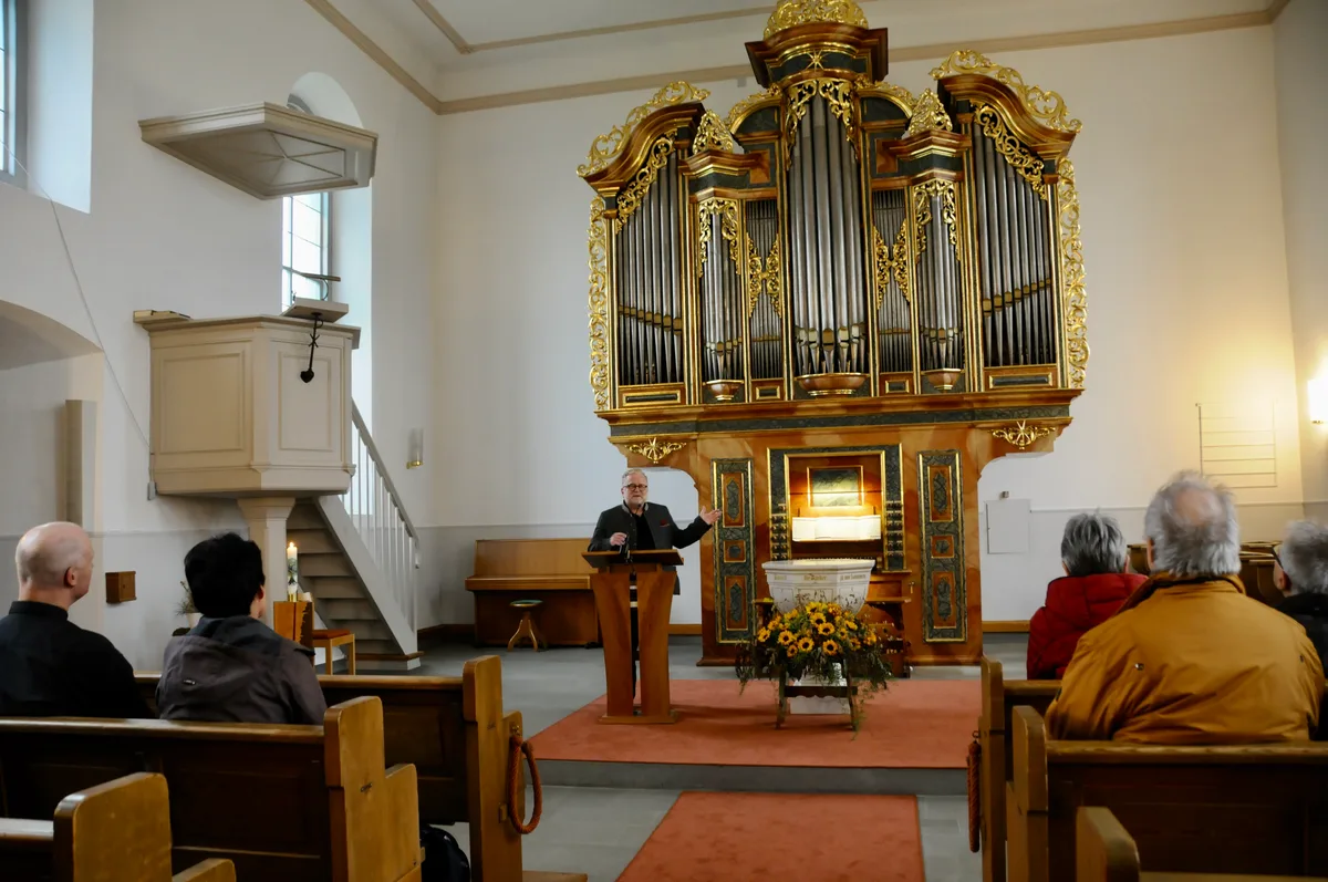 Man sieht einen Pfarrer, der vor Besuchern in der Kirche eine Begrüssungsrede hält.