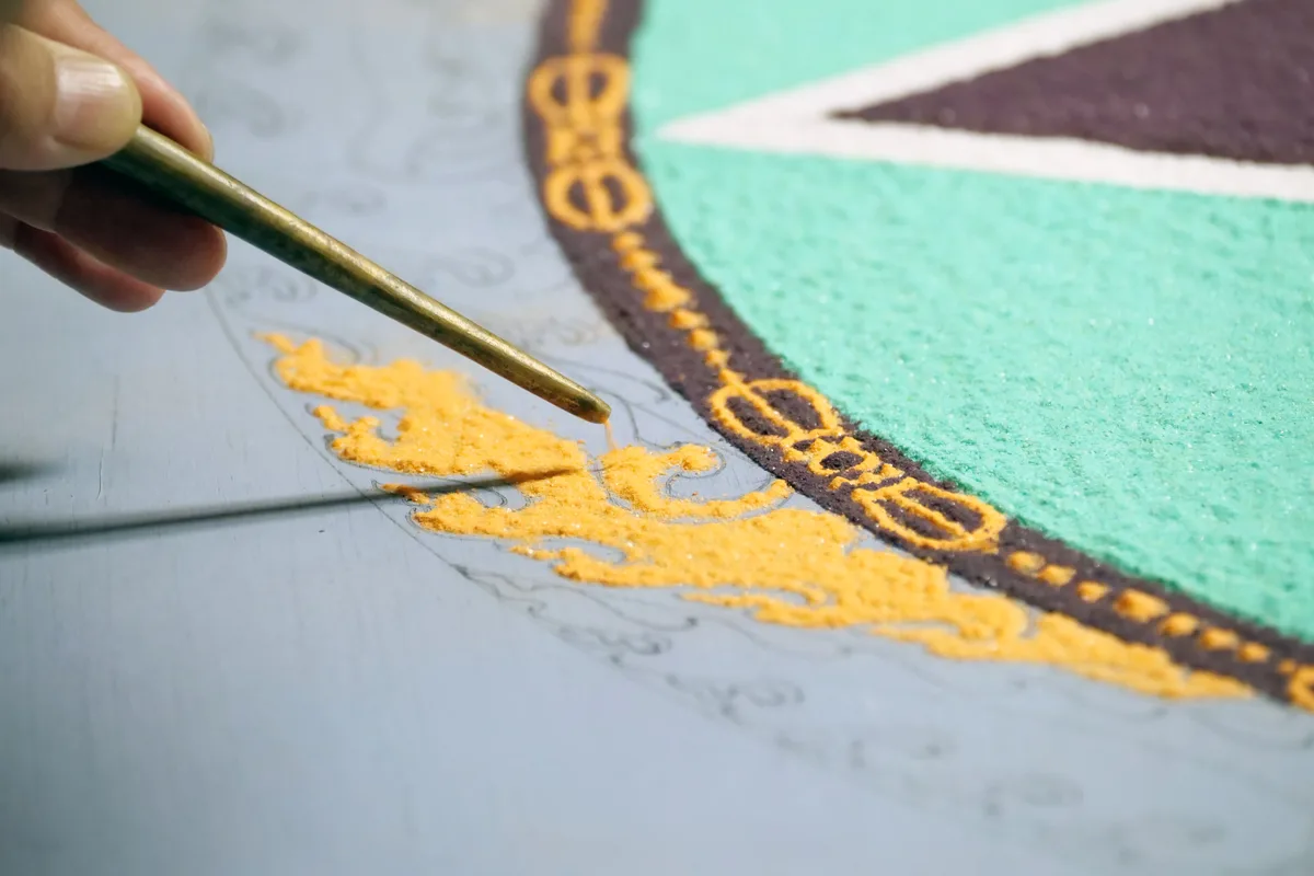 Die mit Bleistift vorgezeichneten Muster werden mit dem farbigen Sand «ausgemalt». Aus der Spitze des «Chakpur-Rohrs» rieselt feiner Sand.