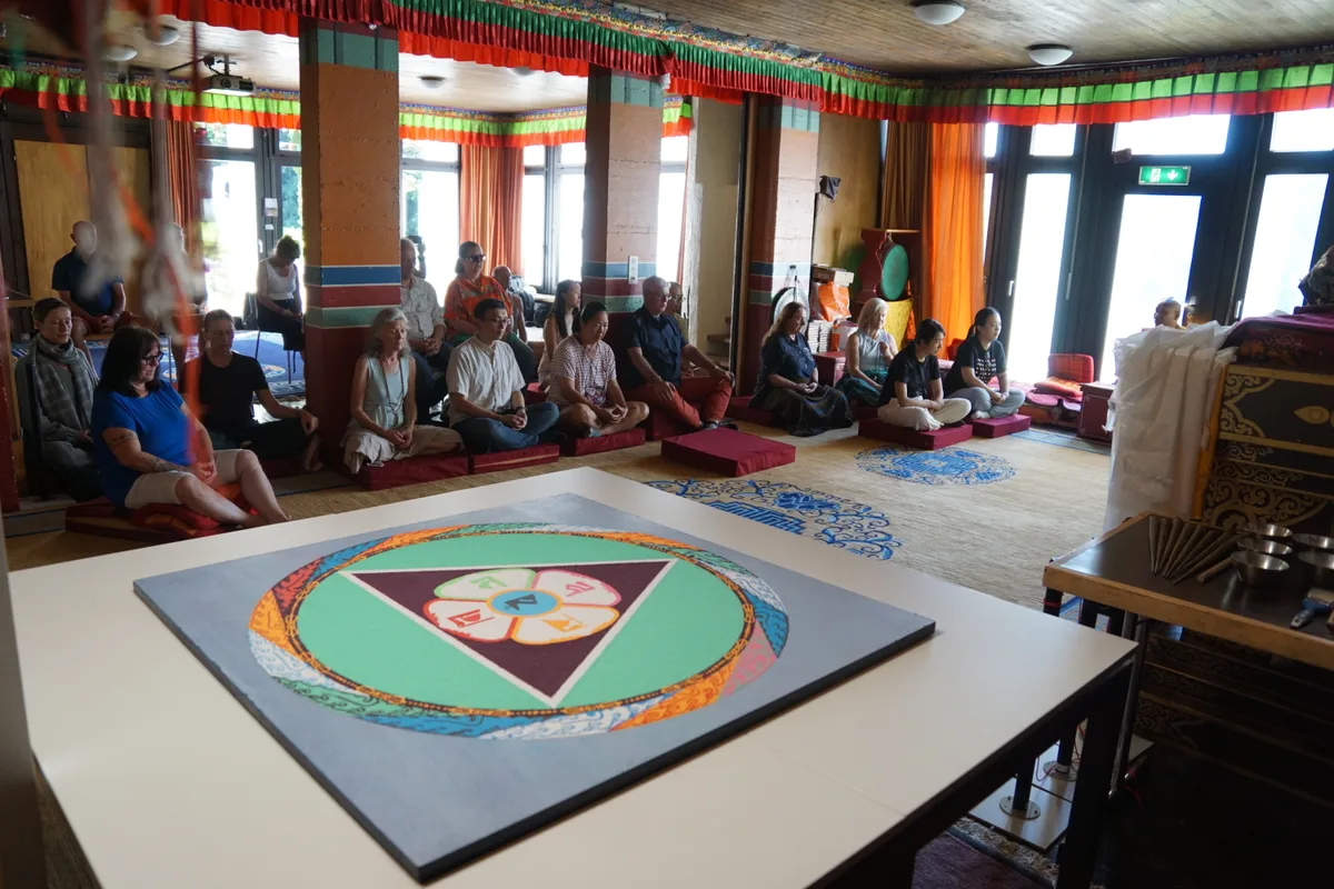 Man sieht Besucherinnen und Besucher im Gebetsraum vor dem Mandala sitzen.