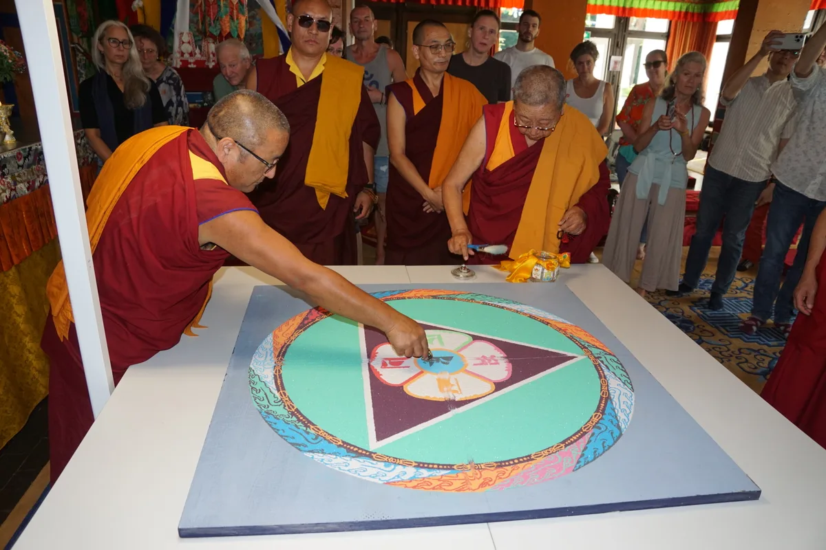 Man sieht buddhistische Mönche, die mit einem spitzen Werkzeug Linien durch ein Sandmandala ziehen.