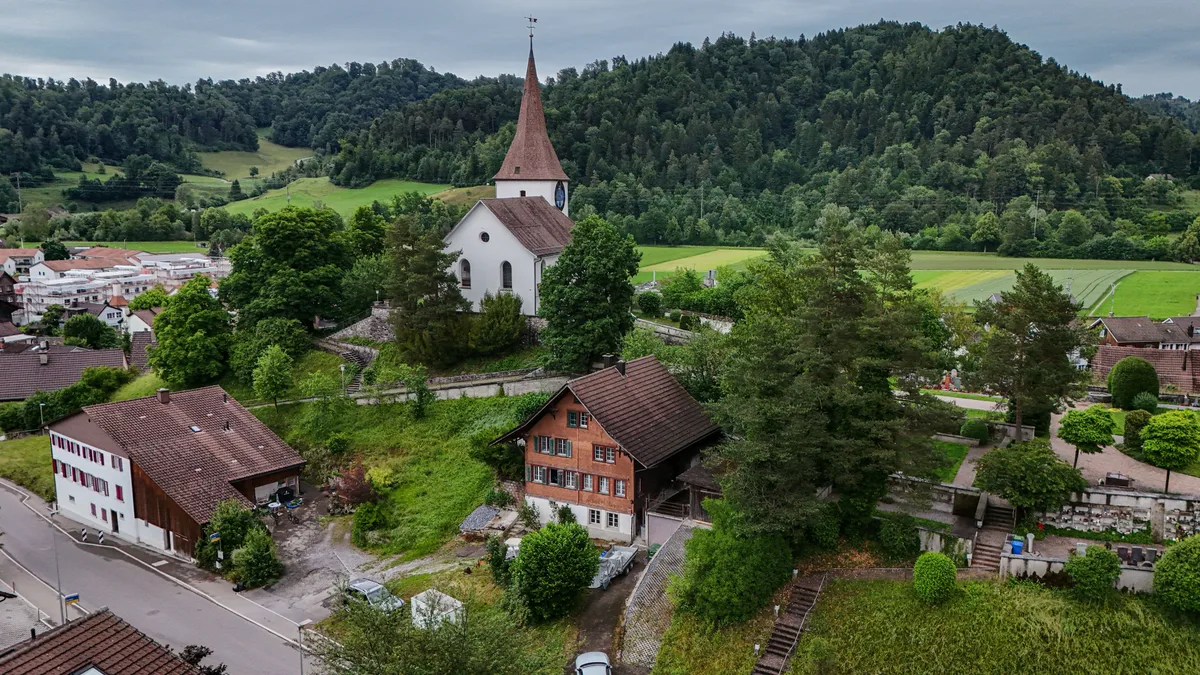 Das Haus liegt direkt zwischen Kirche und Friedhof. Man sieht einen Teil des Dorfs Wila mit der Kirche und dem Friedhof.