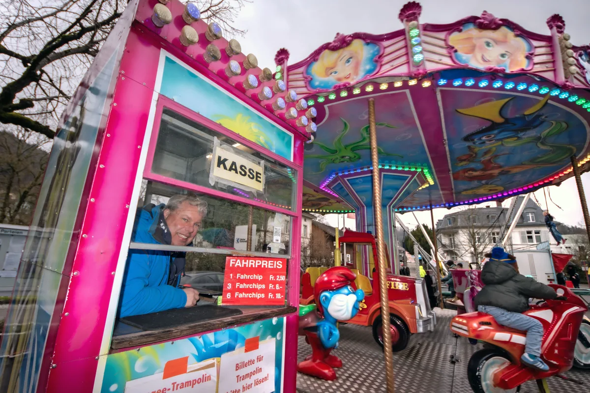 Man sieht einen Mann in einem Kassenhäuschen neben einem Karussell. 