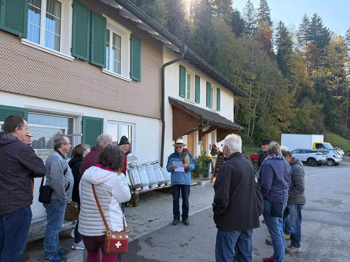 Man sieht eine Gruppe Menschen vor einem Gebäude, vor dem Milchkannen stehen. 