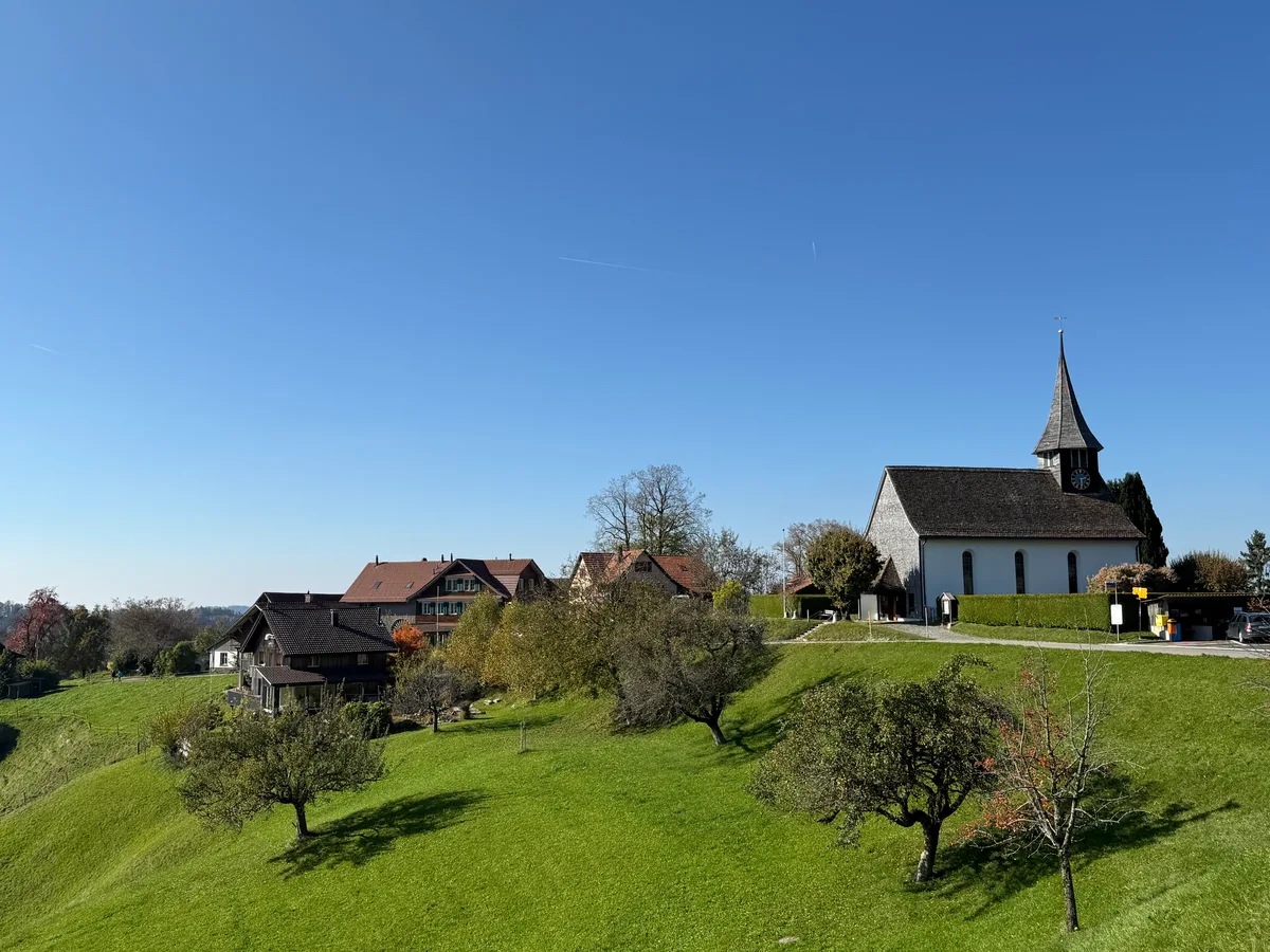 Man sieht das Dorf von Weitem, die Kirche, eine Wiese, Bäume und Wohnhäuser.