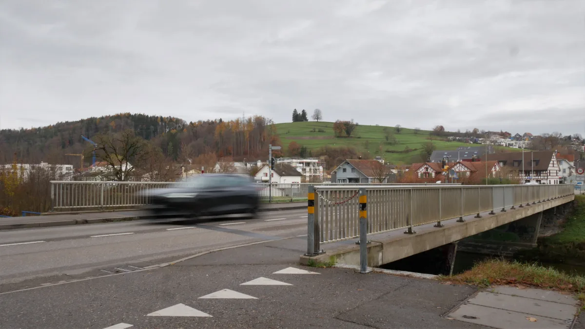 Man sieht eine Strassenbrücke, die die Töss überquert, sowie dahinter Wälder, bei bewölktem Himmel.