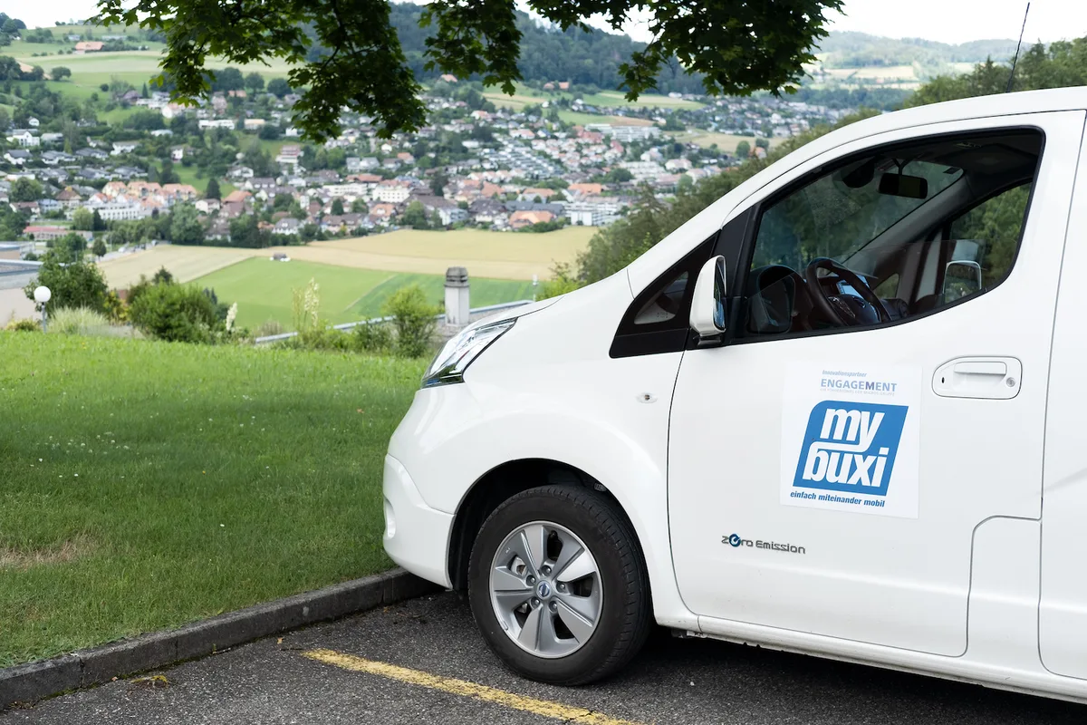 Ein Mybuxi-Ruftaxi steht auf einem Parkplatz. Im Hintergrund Wiesland und ein Dorf.