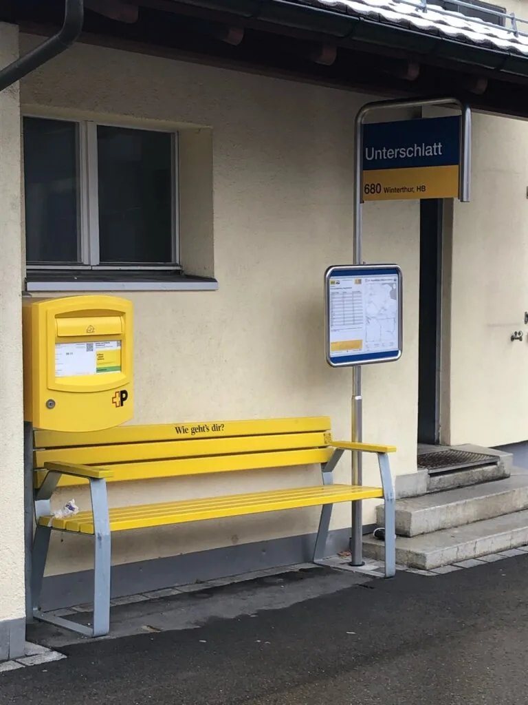 Wer auf das Postauto wartet, kann jetzt in Unterschlatt auf dem «Wie geht’s dir?»-Bänkli Platz nehmen. Man sieht ein gelbes Bänkli an einer Postautohaltestelle.