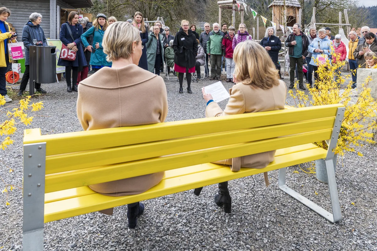 An die Einweihung des neuen Bänkli kamen gut 40 Personen. Zwei Frauen sitzen auf gelber Sitzbank. Im Hintergrund sieht man menschen, die zuhören